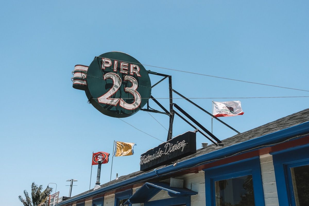 The sign for Pier 23 Cafe Restaurant with a blue sky in the background, with a 49ers flag, a warriors flag, and California state flag visible.