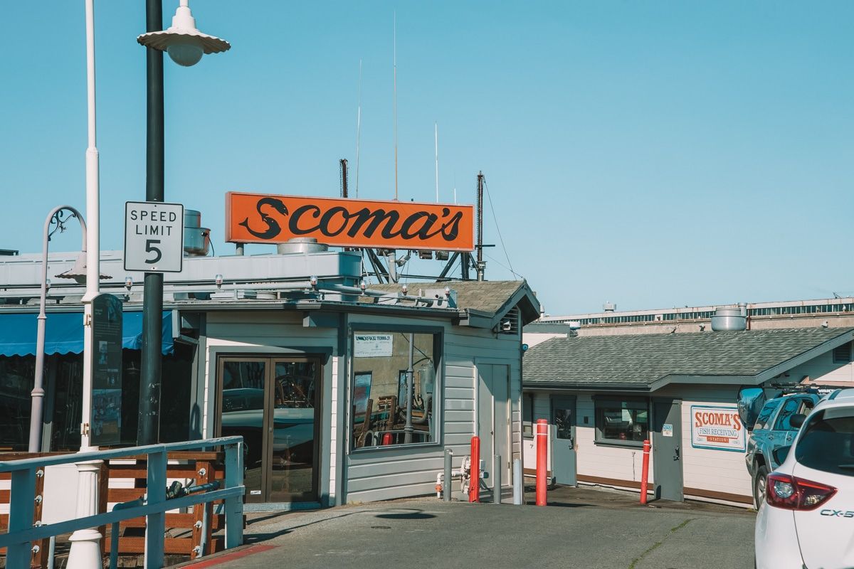 Scoma's restaurant, a small building with wood siding, with a large orange and black sign on the roof, and a cloudless blue sky behind.