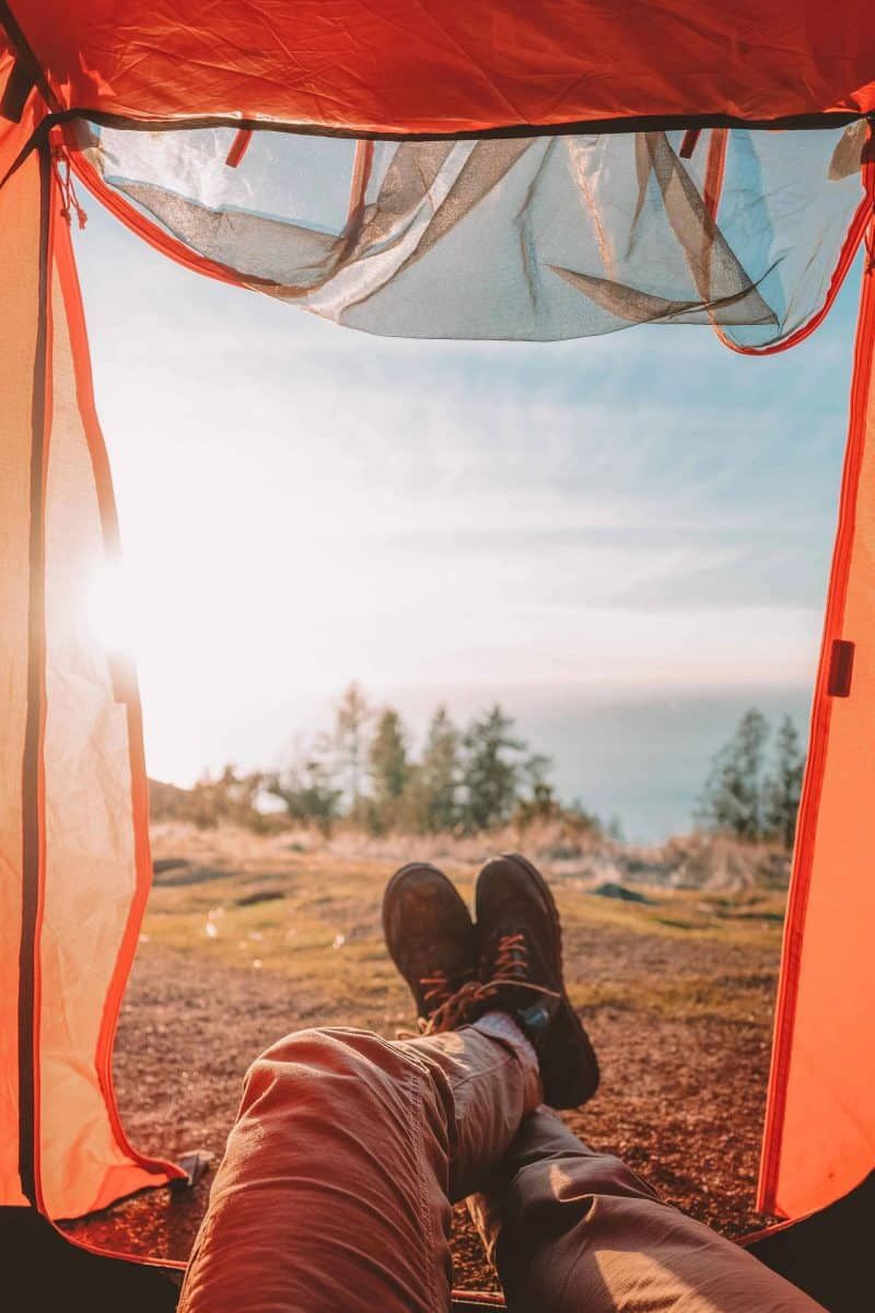 Point of view of a person's legs crossed as they lounge in the open doorway of an orange tent, with trees on the horizon.