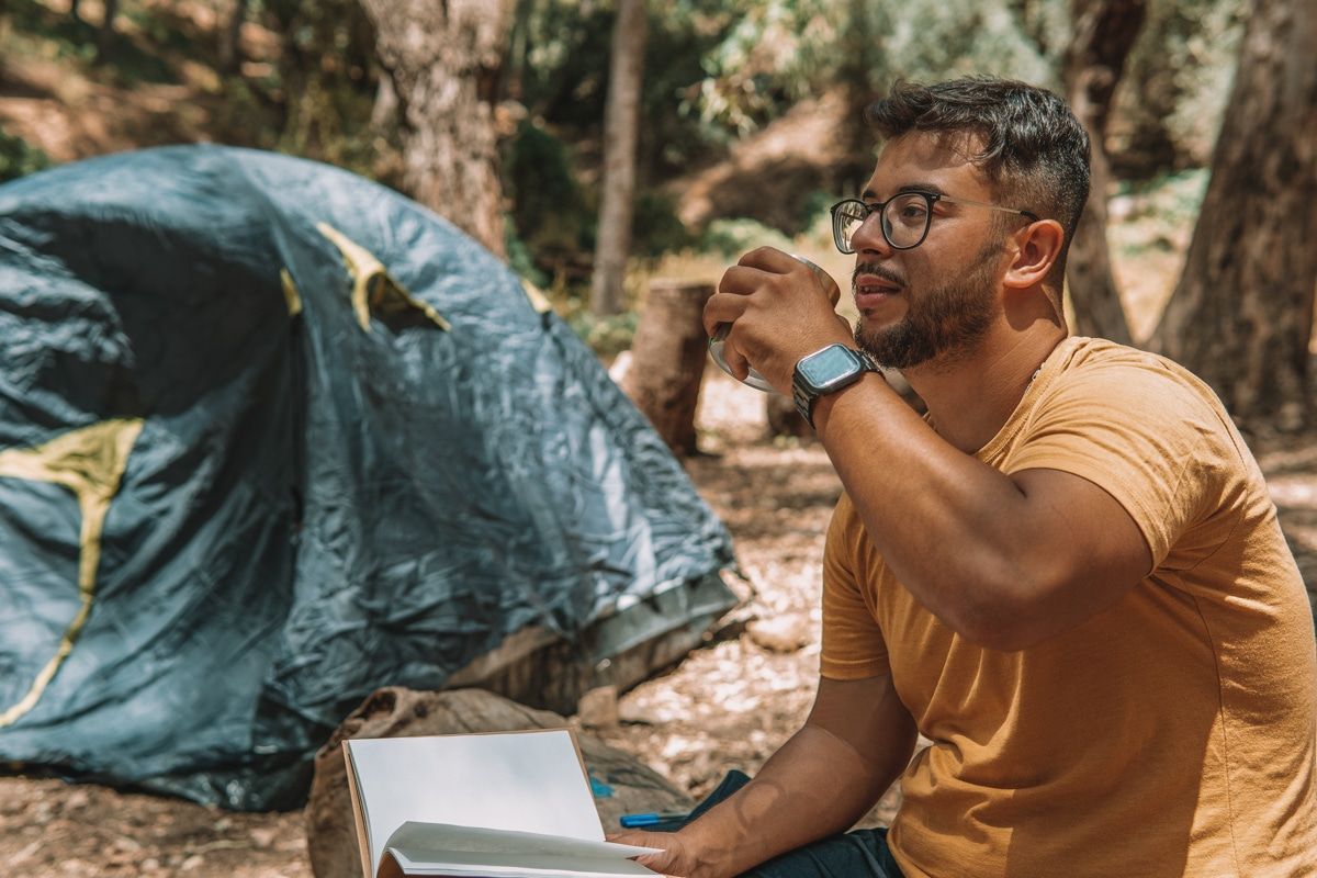A man in glasses and a yellow shirt sits at a campsite, holds an open book in one hand, and raises a mug to his lips with the other.