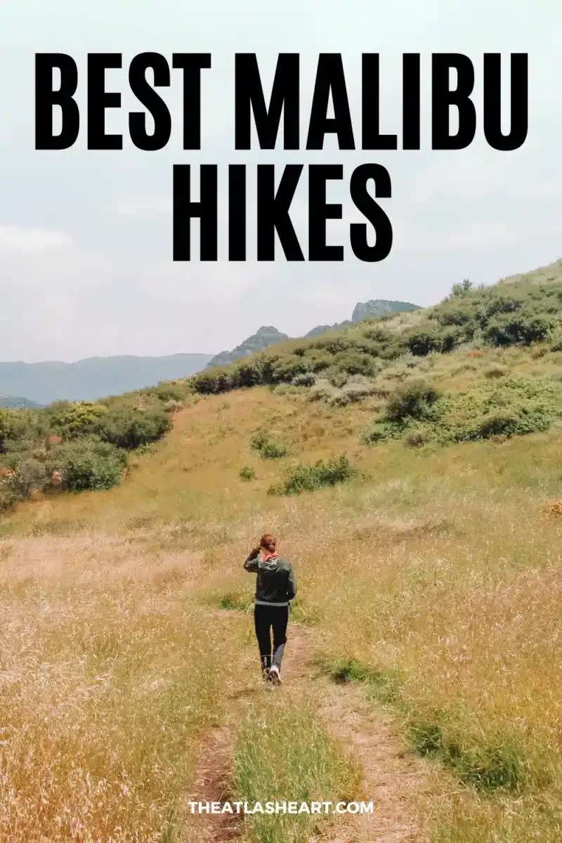 A woman seen from behind while hiking on a trail through a grassy valley, with mountains and a partly cloudy sky in the background, and the text overlay, "Best Malibu Hikes."