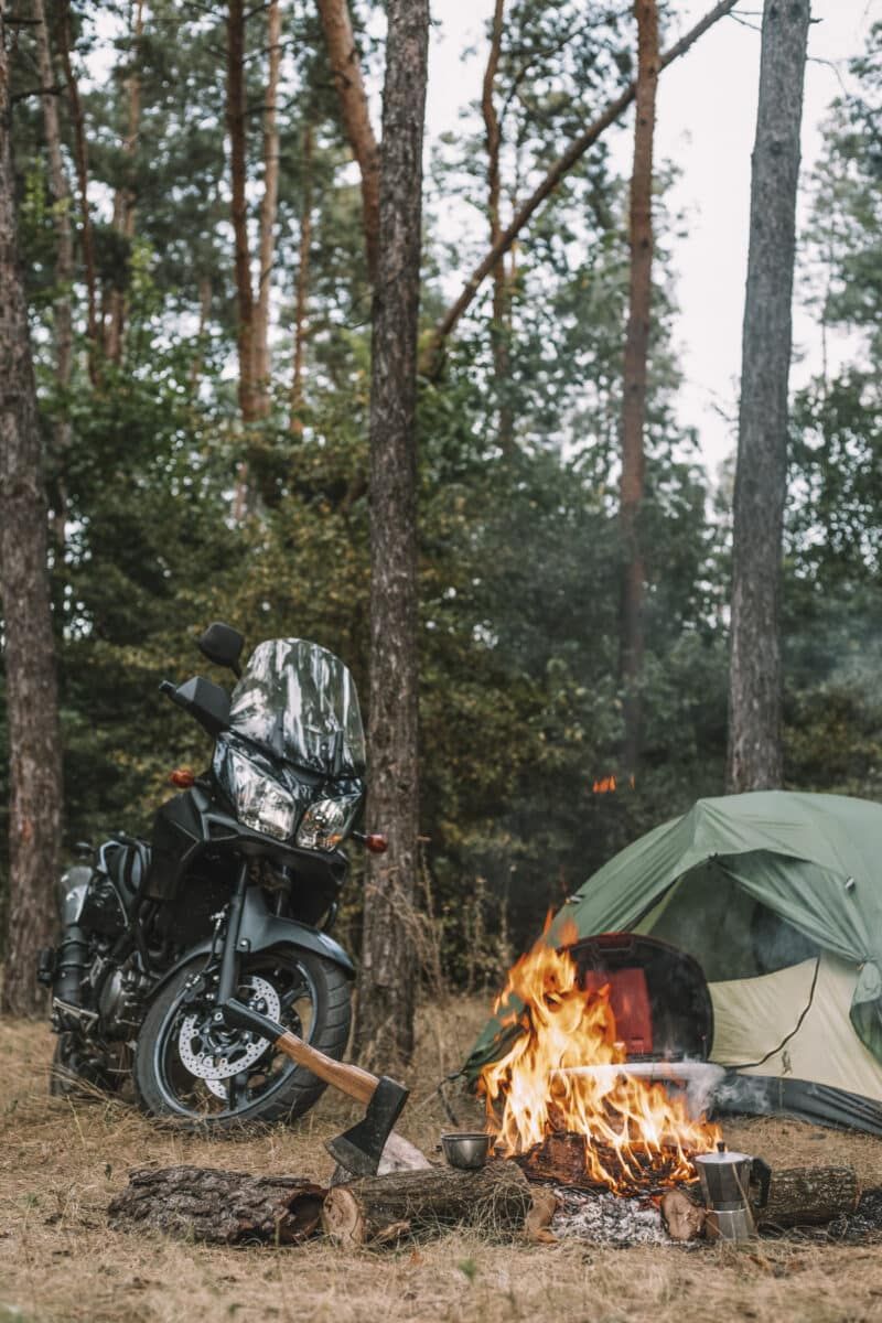 A roaring campfire with an ax beside it, and in the background a motorcycle and one of the best motorcycle tents.