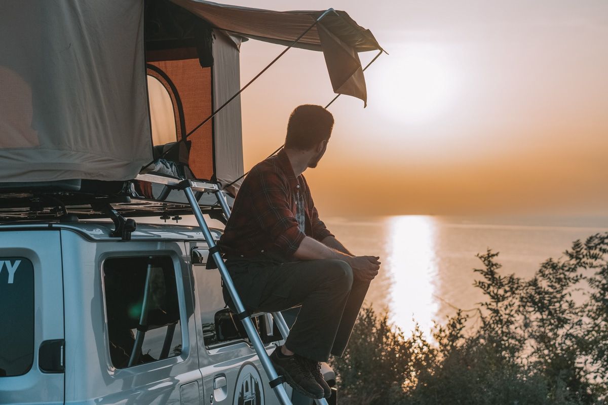 A man sitting on the ladder of a rooftop SUV tent, looking out over a body of water at sunset.