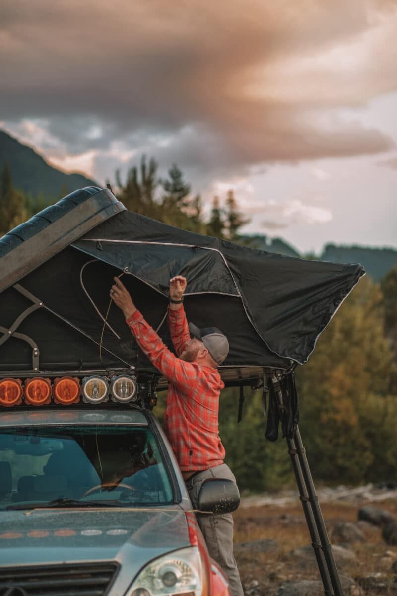 A man wearing a red flannel setting one of the best SUV tents on the roof of his truck, with trees and a cloudy sky in the background.