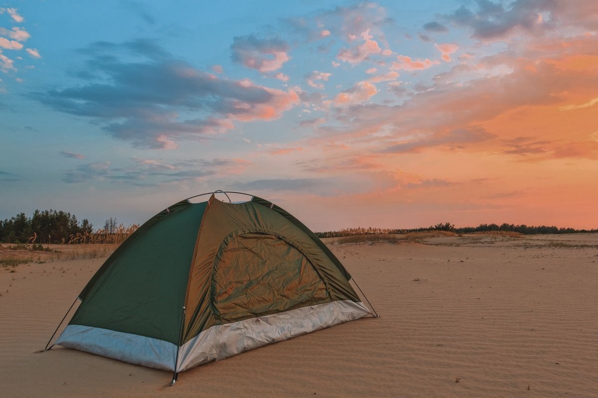 A small, green, two-person tent on a beach at sunset, with a pink and blue sky in the background.