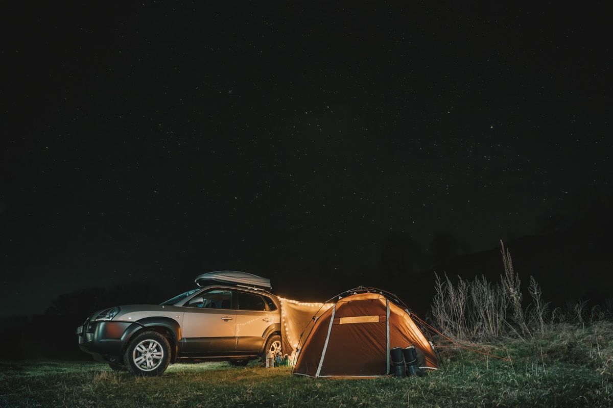 A nighttime scene of 
a small hatchback SUV tent pitched at a campsite next to a silver SUV, illuminated by string lights.