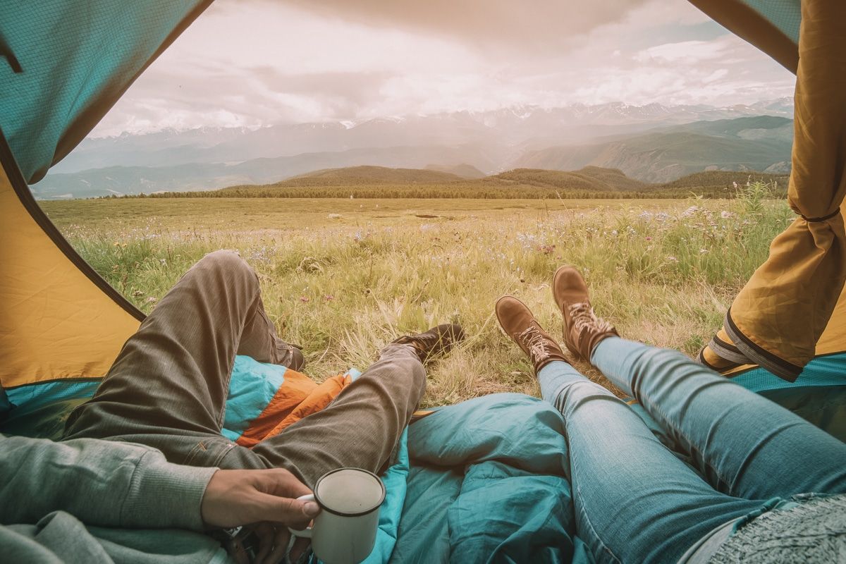 POV of two people lounging with their legs sticking out of the doorway of a small tent, looking out on a grassy landscape.