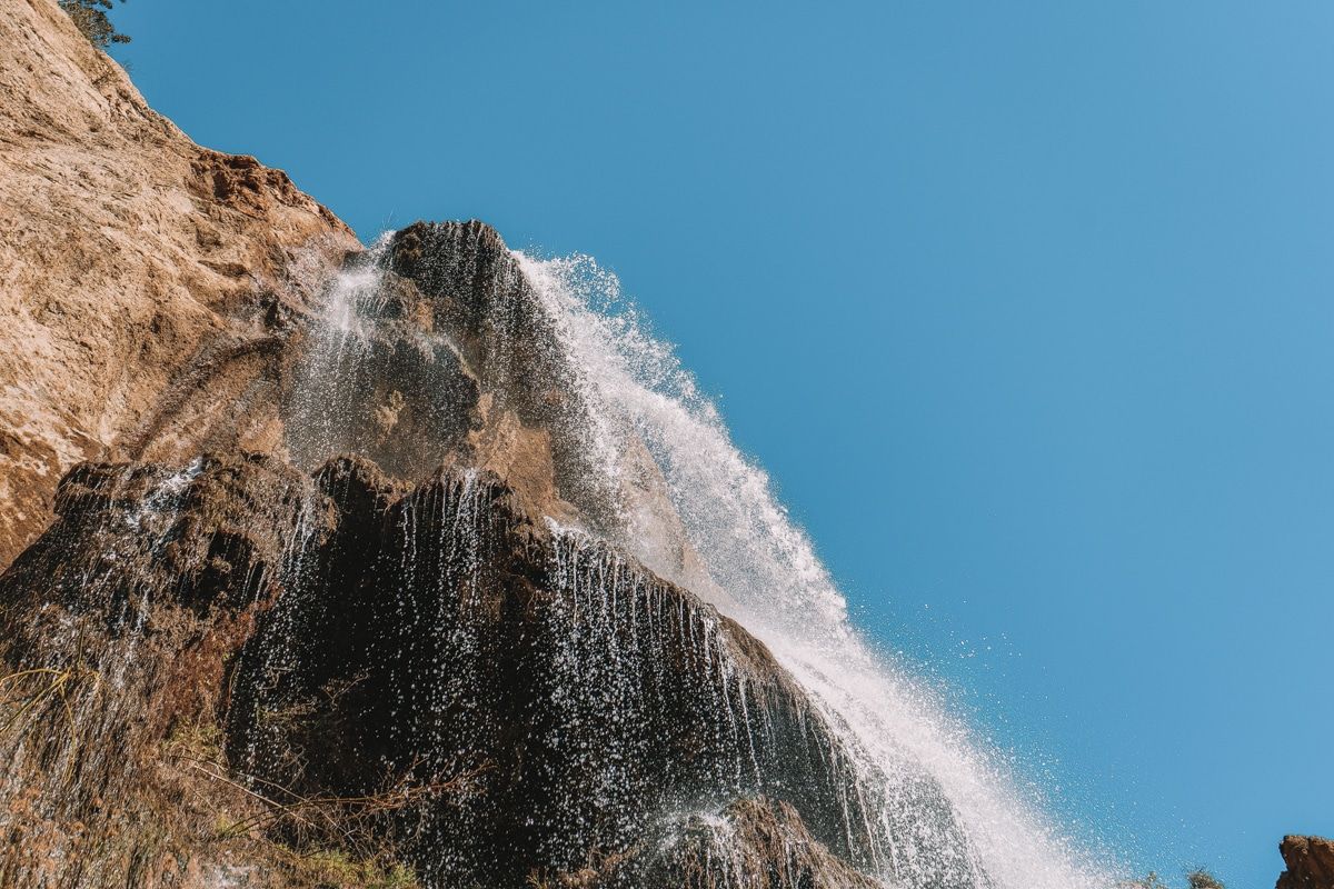 A close-up view from below Escondido Falls, a waterfall splashing over brown rocks with a clear blue sky in the background.