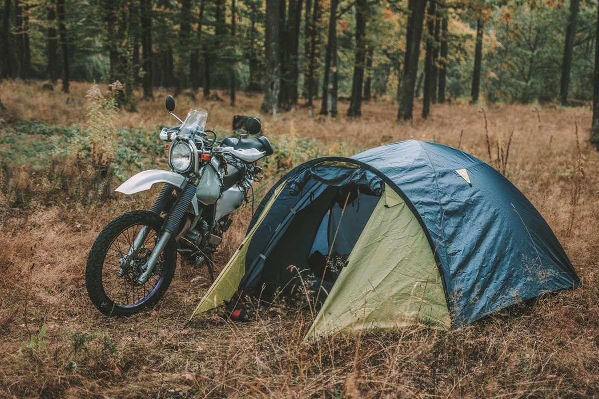 A motorcycle tent pitched in a grassy campsite beside a motorcycle, with trees in the background.