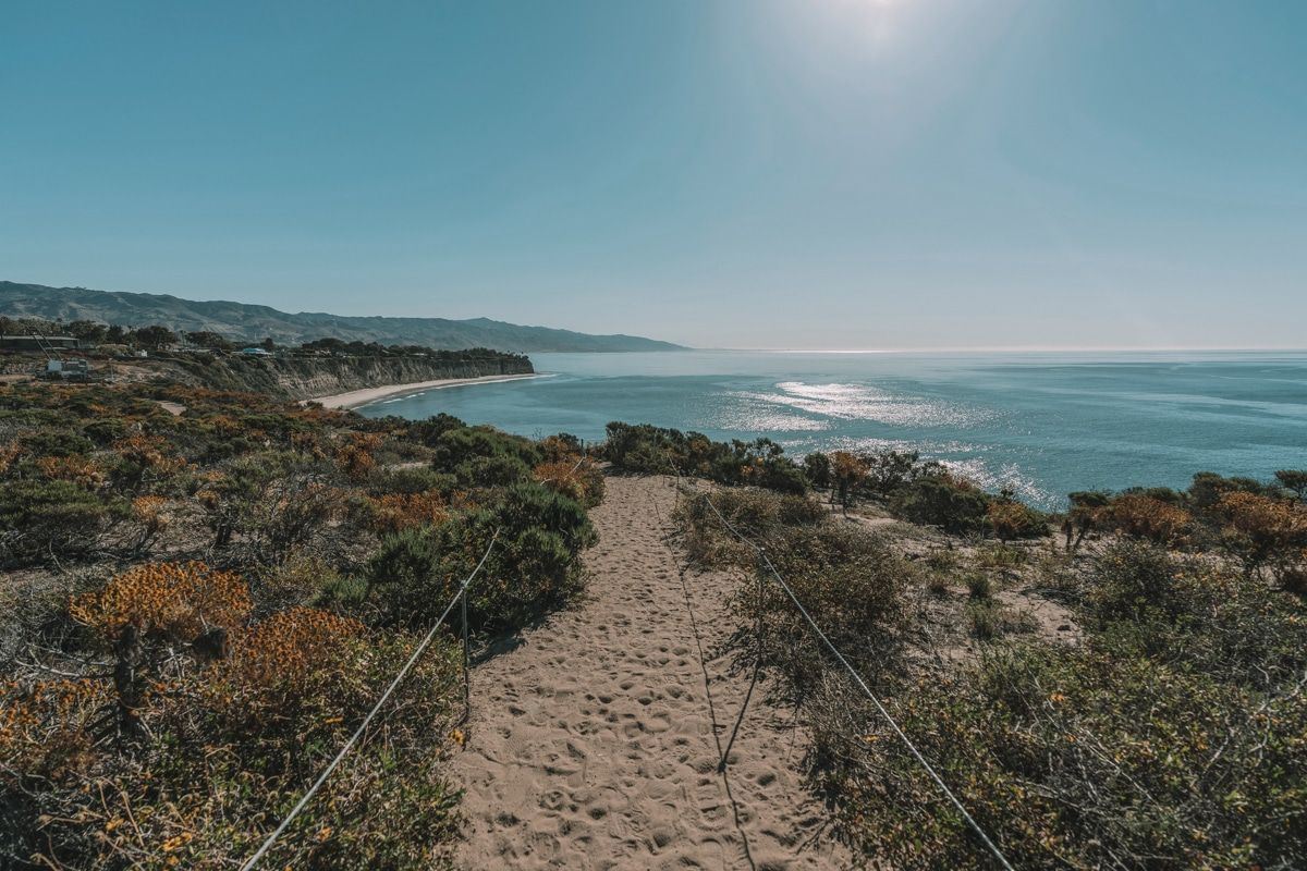  Point Dume Cove Trail, a sandy path lined by scrub brush, with the sun reflecting off the ocean in the distance.