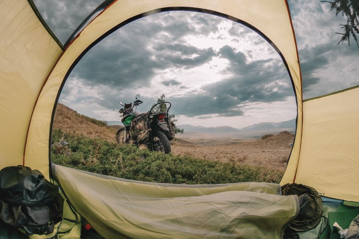 The view of a motorcycle parked in a grassy landscape seen from inside a tent.