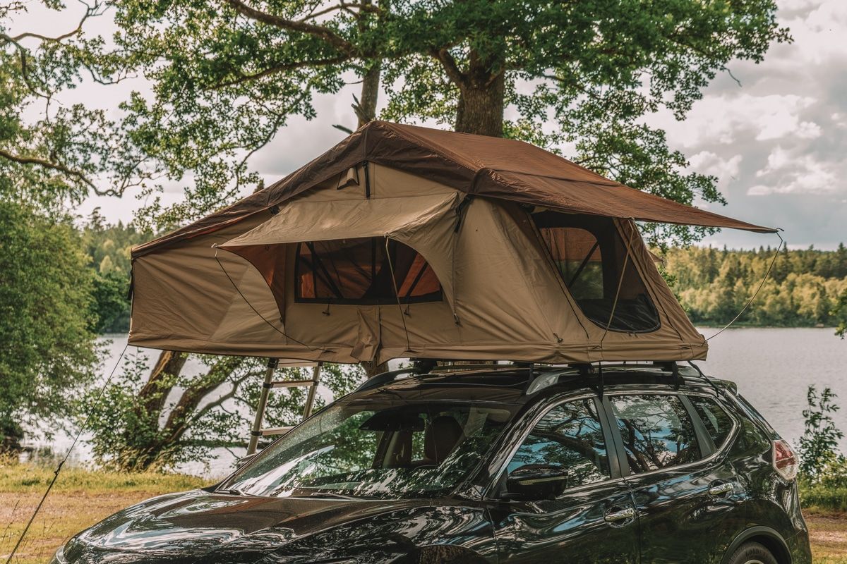 A brown SUV tent on the roof of a black SUV, with trees and a lake in the background.