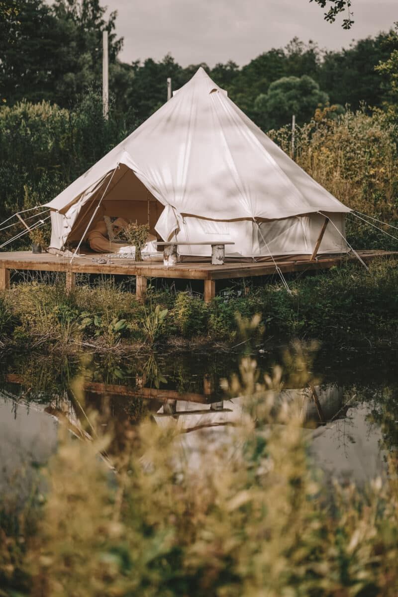 One of the best glamping tents, a canvas bell tent, on a wooden platform beside a pond surrounded by vegetation.