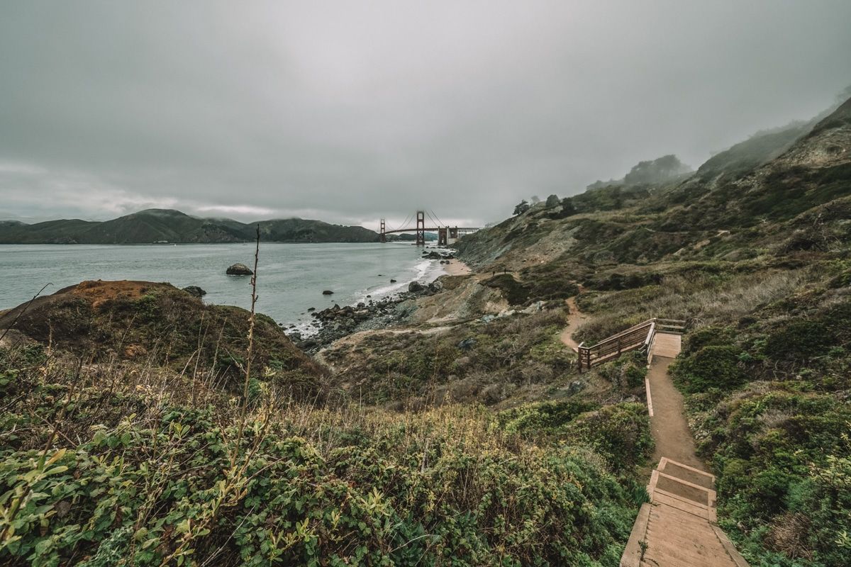 View of a trail through low scrub brush leading down toward the water, with the Golden Gate Bridge and a cloudy sky in the distance.
