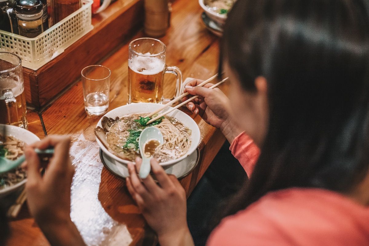 A view over the shoulder of a woman enjoying a bowl of ramen and a glass of beer at a restaurant counter.