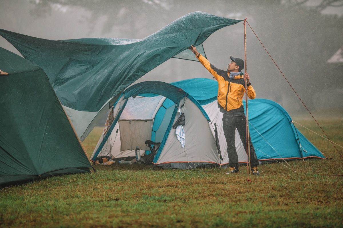 A man in a yellow windbreaker attempts to place a green rain fly over his blue tent as it blows in the wind.