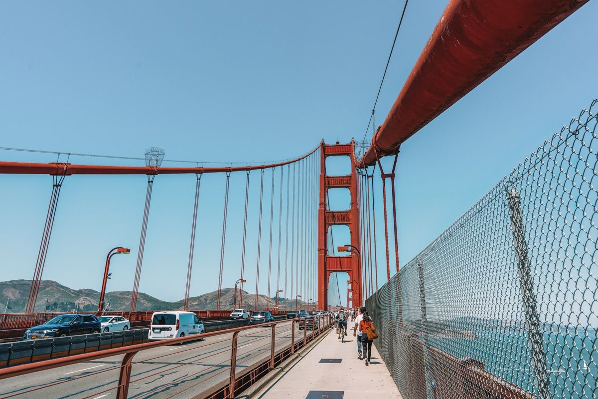 People seen from behind as they walk on the Golden Gate Bridge pedestrian path, with the tower and cables in forced perspective.