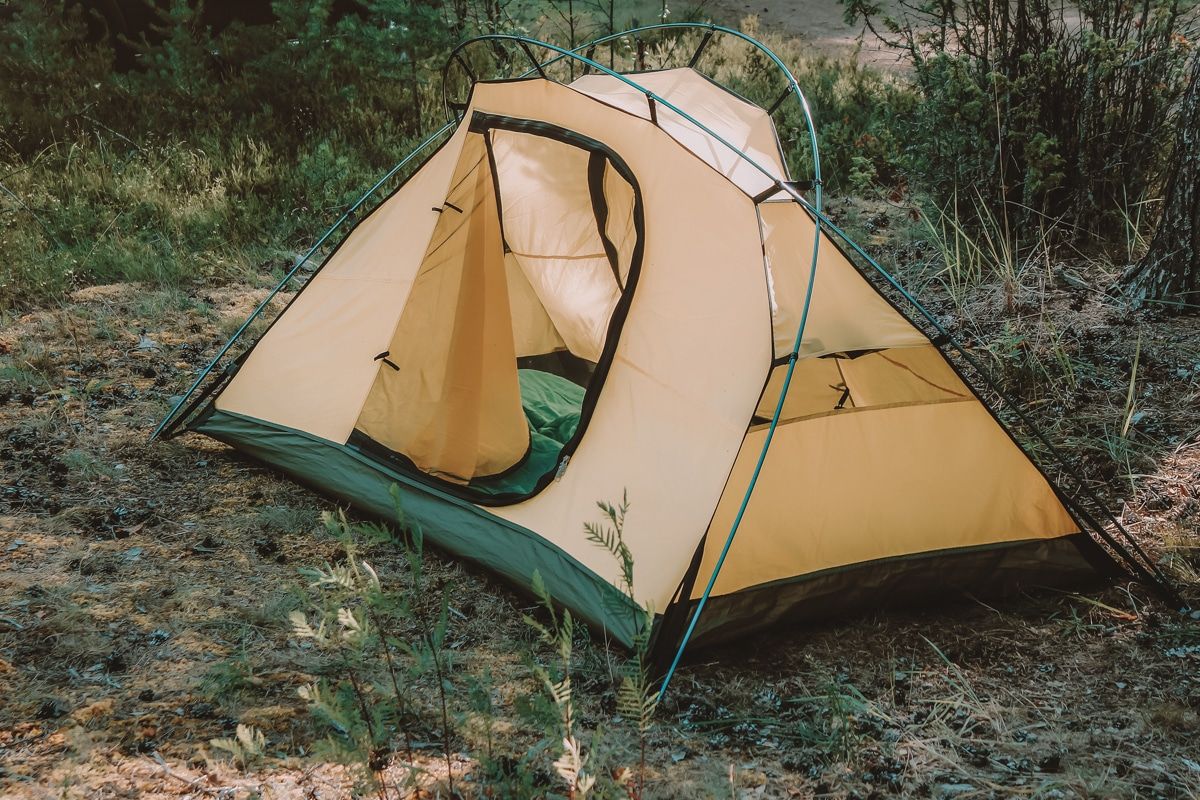 A tan one-person tent with green trim pitched on a mossy patch of ground with vegetation in the background.