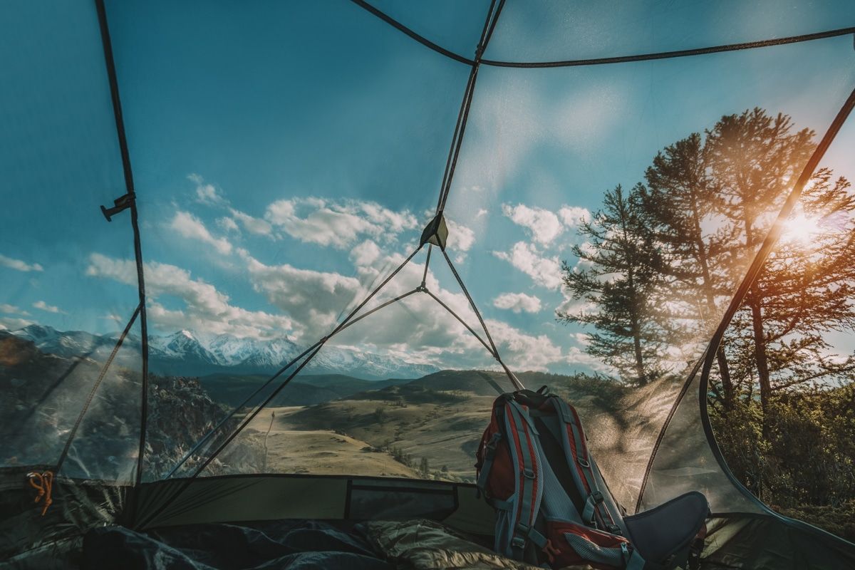 View of a wide-open, hilly landscape and a blue sky with a few clouds seen through a mesh tent screen room, with a backpack in the foreground inside the tent.