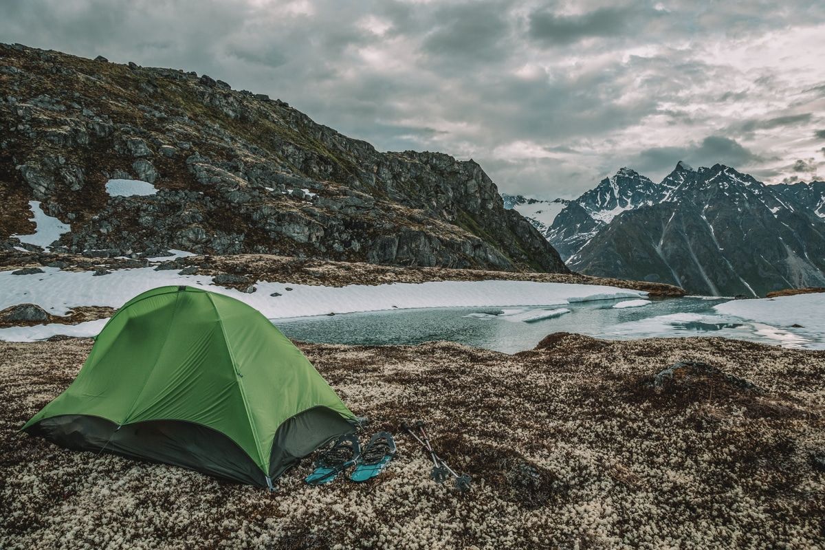 A small tent with a green rain fly set up beside an icy pond in a wintry mountain landscape.