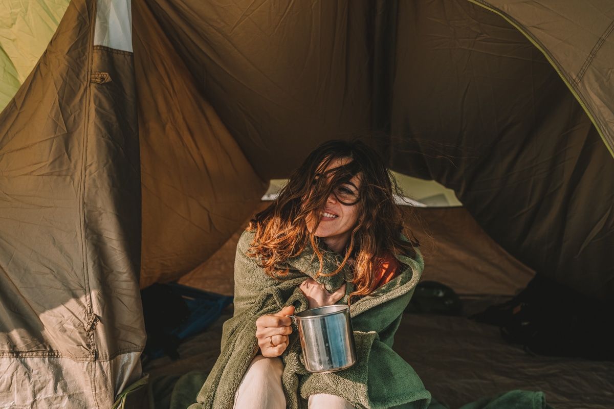 A young woman with brown hair hugs a blanket around herself, smiles, and sips coffee from a tin mug in the doorway of a brown tent as the wind blows her hair over her face.