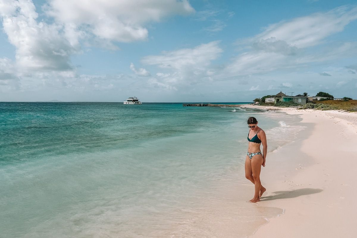 A woman in a bikini looks down at her feet while standing on a white-sand beach at the shore of a calm, turquoise ocean, with a partly cloudy, blue sky above.
