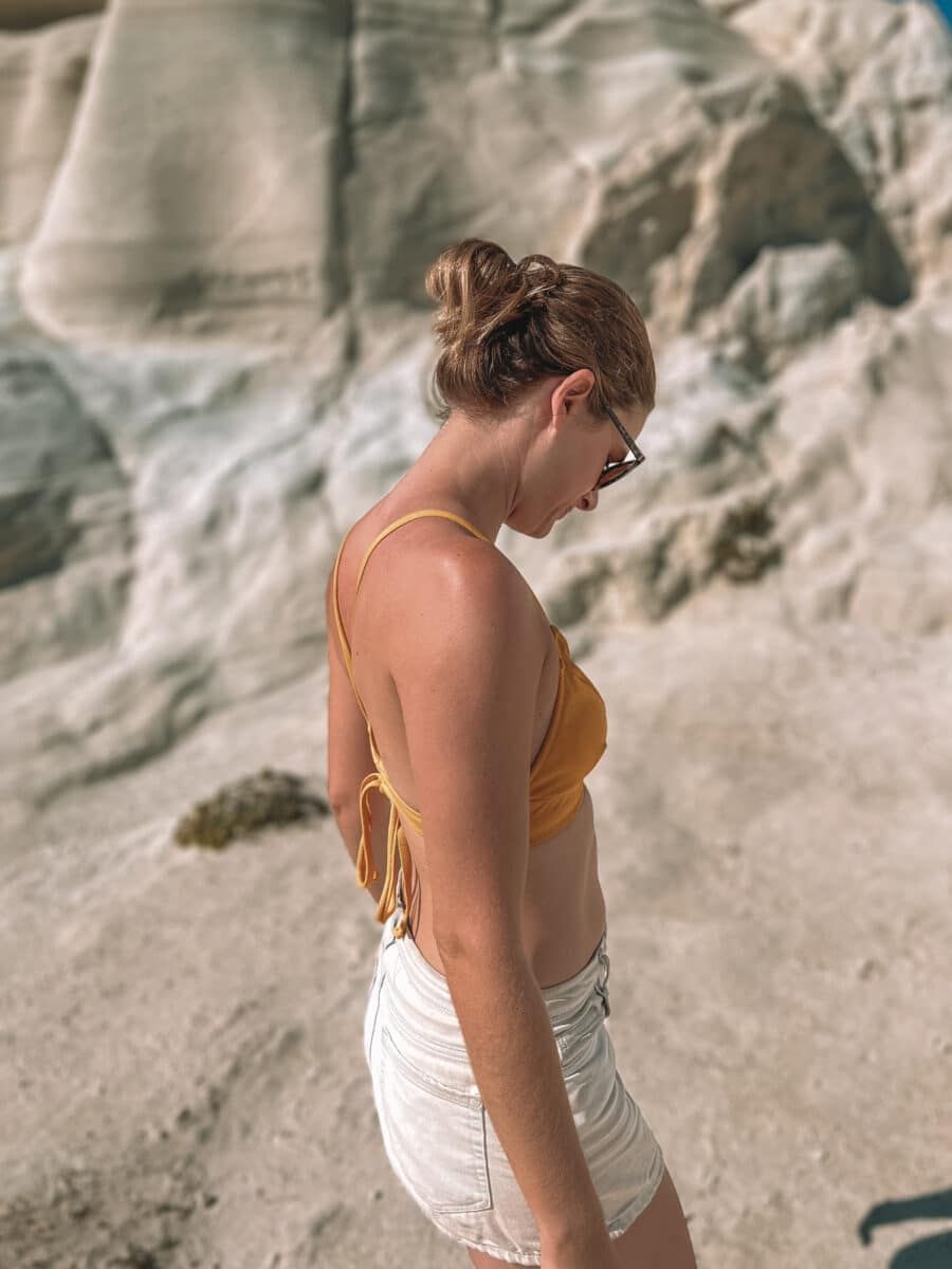 Close-up of a woman in profile, wearing a yellow bikini top and white shorts, looking down, with white sandstone rock formations and a clear blue sky in the background.