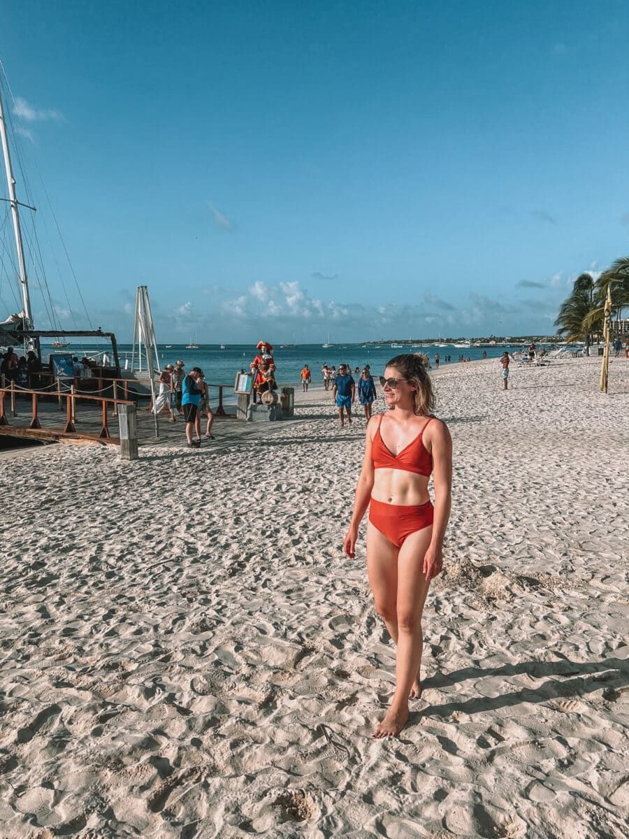 A woman wearing sunglasses and a rust red, high-waisted bikini walking toward the camera on a tropical beach with sparse crowds in the background.