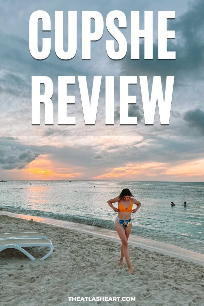 A woman in a yellow bikini top and blue leaf-printed bottoms adjusts her top  and looks down at her feet while walking on a tropical beach during a slightly overcast sunset, with the text overlay, "Cupshe Review."