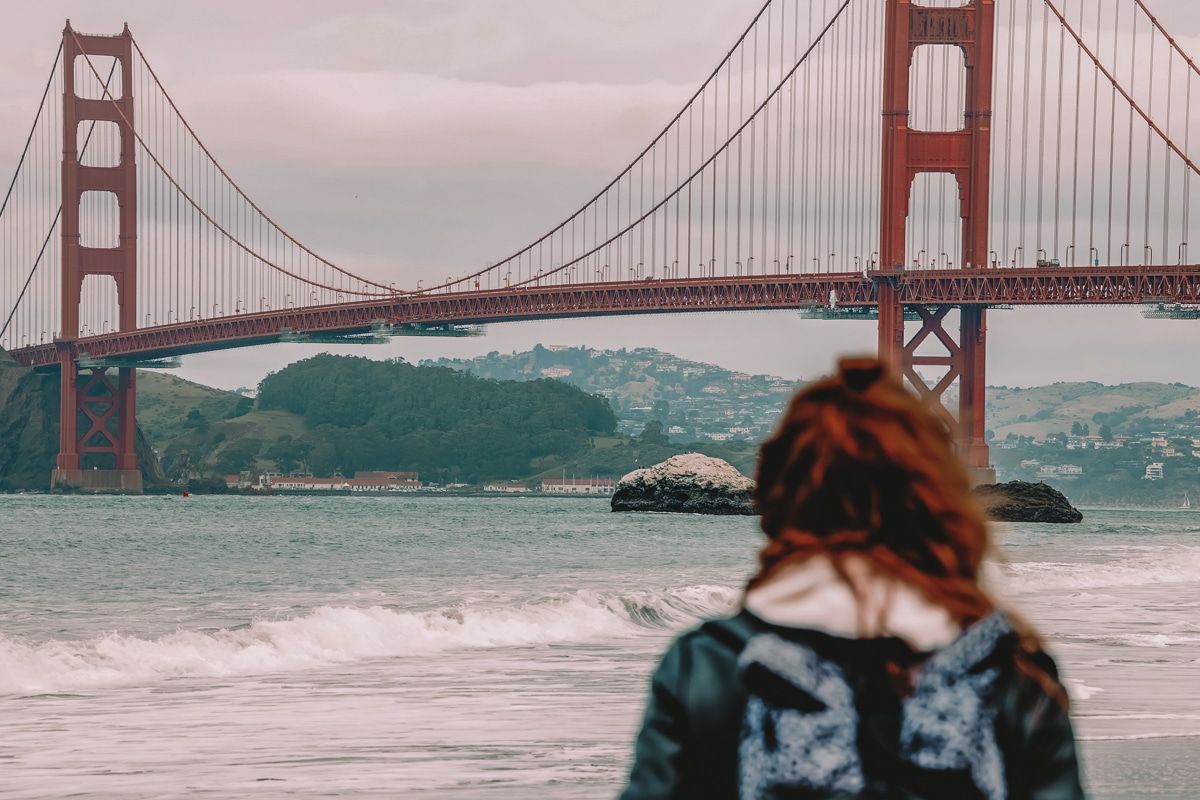 A red-haired woman seen from behind as she looks out over the water at the Golden Gate Bridge on a cloudy day.