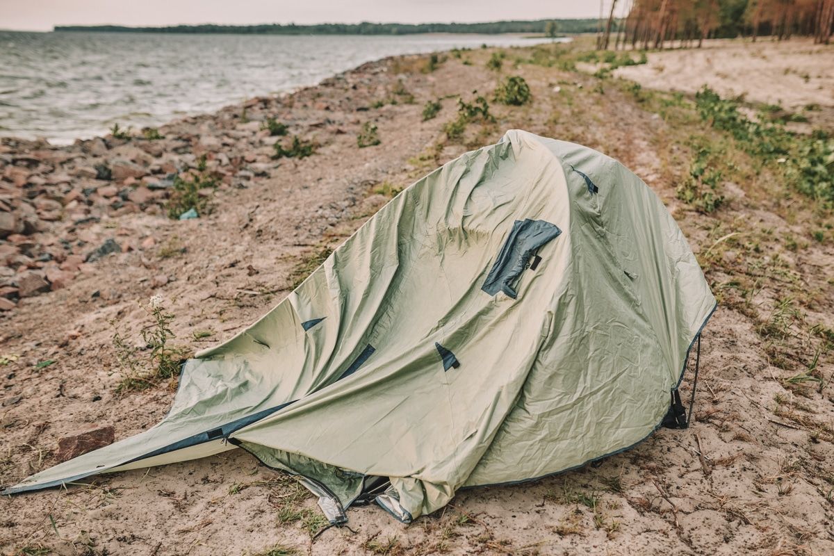 A windblown, partially collapsed tent draped in a light green rainfly, pitched on the banks of a river.