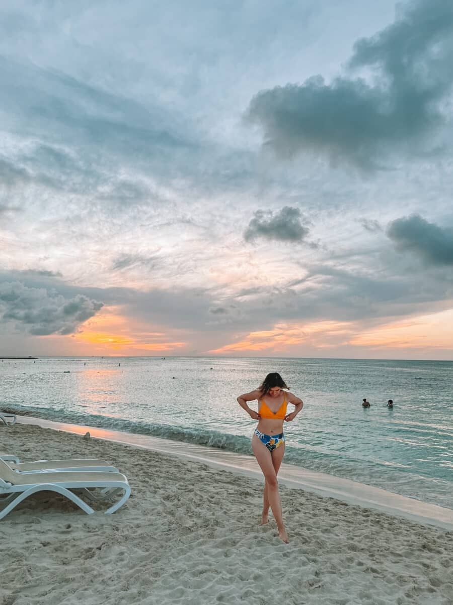 A woman in a yellow bikini top and blue leaf-printed bottoms adjusts her top  and looks down at her feet while walking toward the camera on a tropical beach during a slightly overcast sunset.