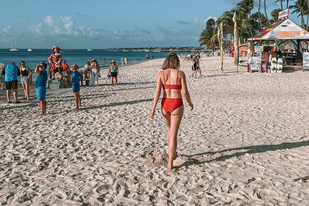 A woman wearing a rust red, high-waisted bikini walking away from the camera on a tropical beach with sparse crowds in the background.