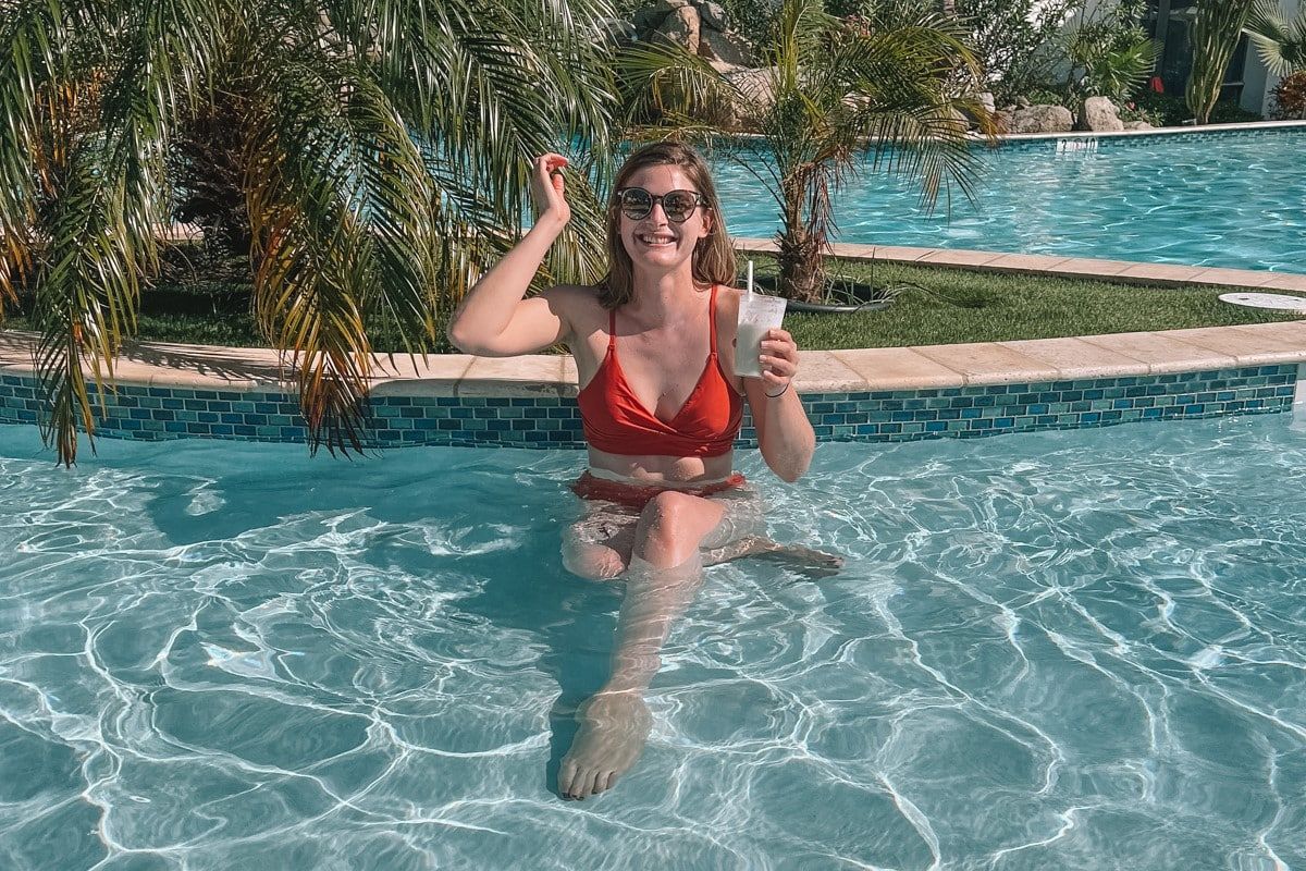 A woman wearing sunglasses and a rust red, high-waisted bikini grins while holding a cocktail and sitting in a pool, with palm trees in the background.