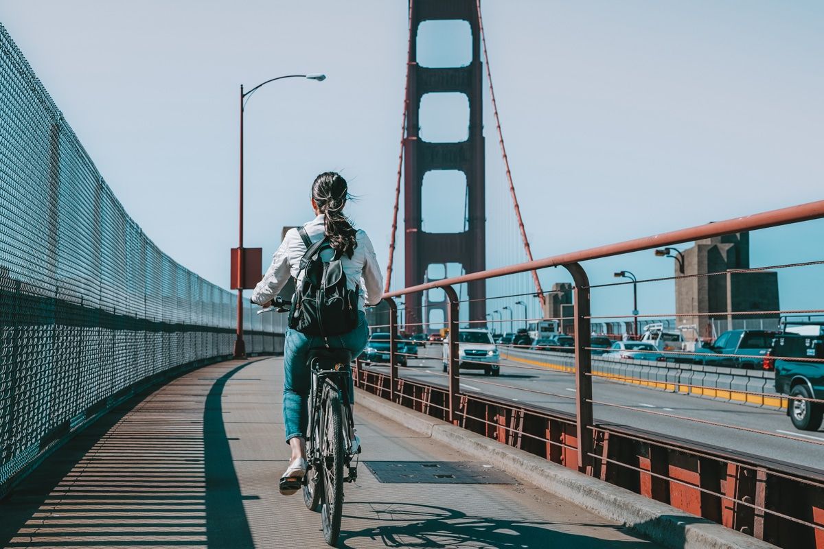 A woman with blakc hair tied in a ponytail seen from behind as she bikes across the Golden Gate Bridge.