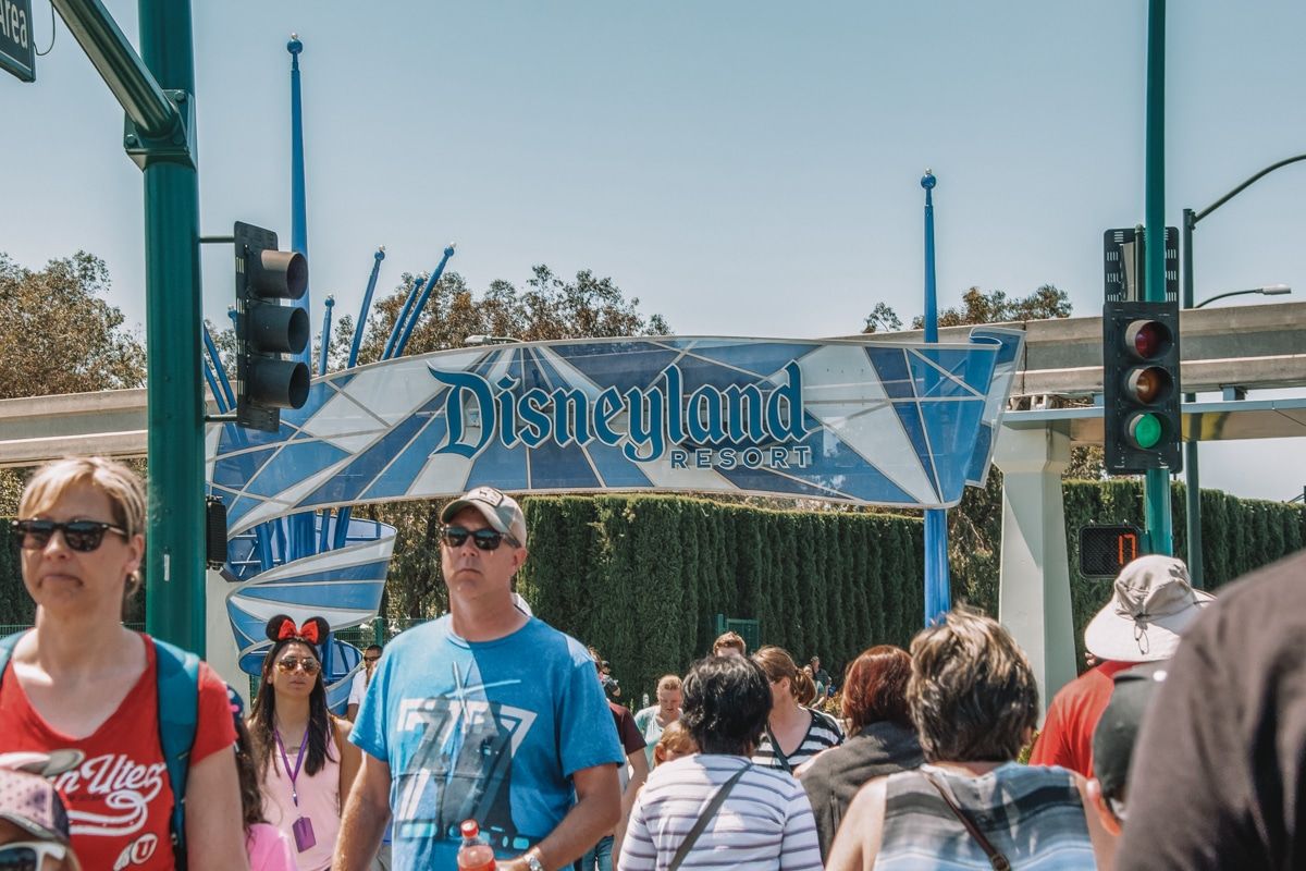 A crowd of people moving through Disneyland on a sunny day, with a blue Disneyland sign in the background.