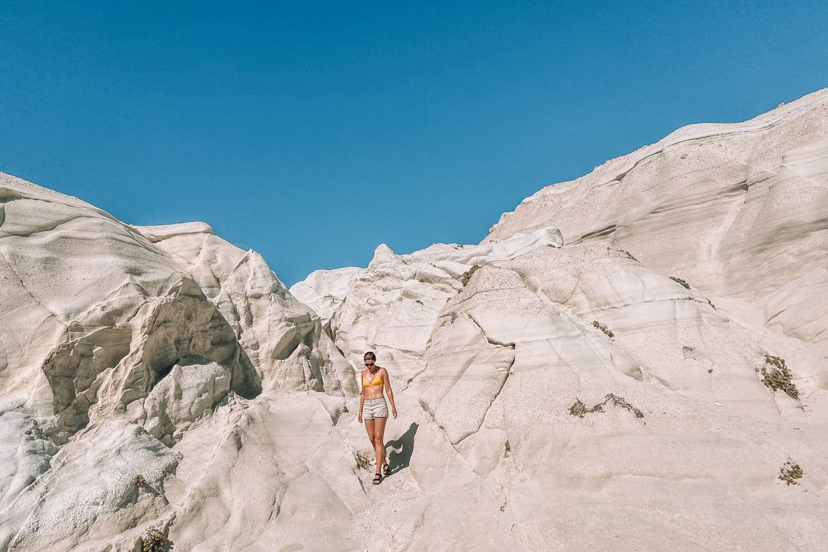 A woman wearing a yellow bikini top and white shorts picks her way over white sandstone rock formations, with a clear blue sky in the background.