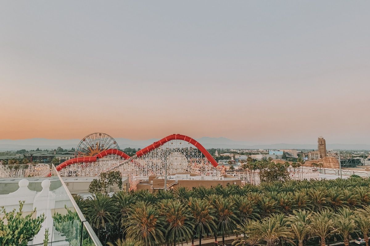 A view of Disneyland from afar and slightly above, showing roller coasters, palm trees, and a light sunset in the background.