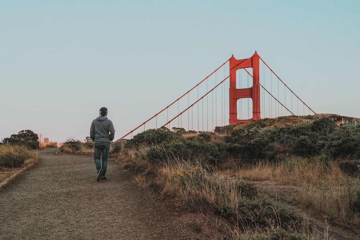 A man in a grey sweatshirt walking along a path with a red tower of the Golden Gate bridge peeking over the ridge.