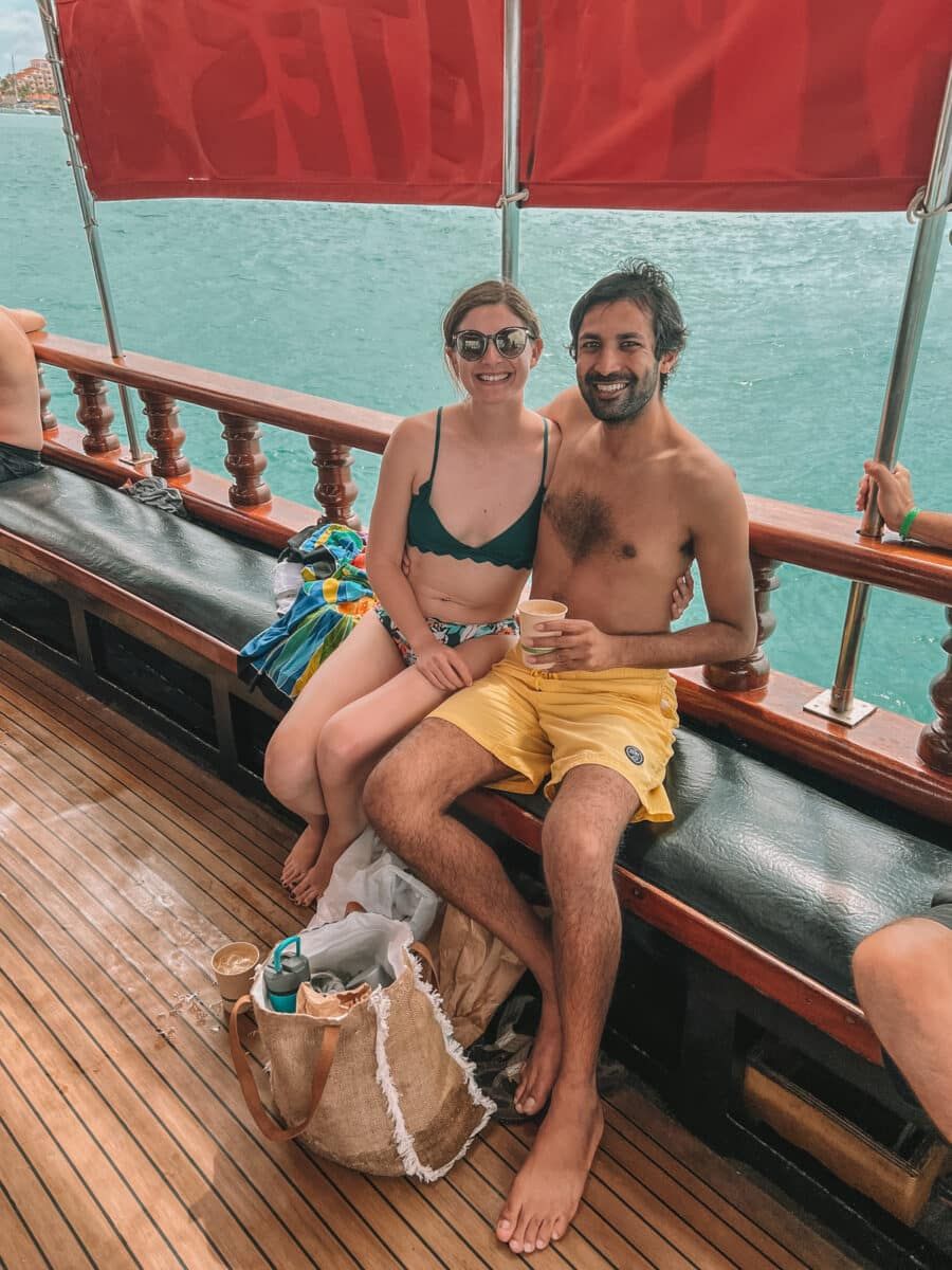 A woman wearing sunglasses, a green, scalloped-edge V-Neck bikini top, and a tropical-printed bikini bottom smiles with her arm around a man in yellow swim trunks as they sit side-by-side by the rail of a ship, with aqua ocean waters in the background.