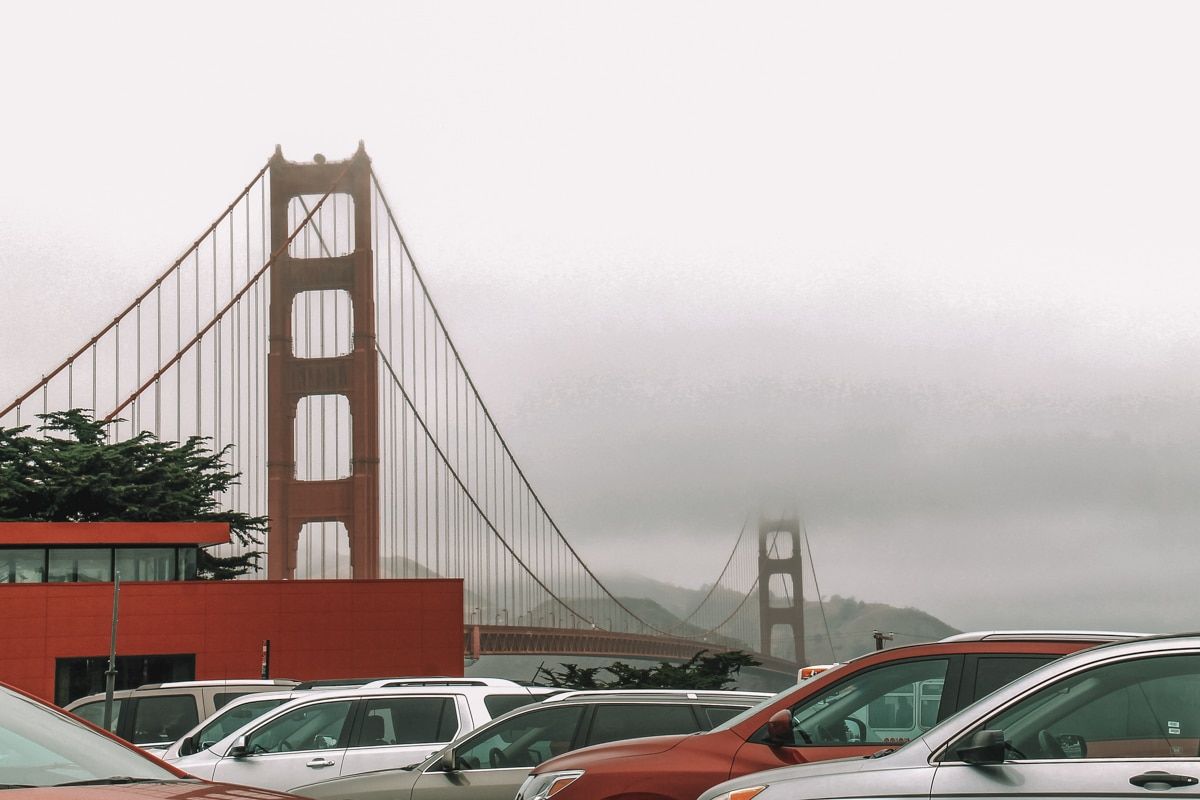 The tops of several cars in a parking lot, with the Golden Gate Bridge visible in the background against a foggy sky.