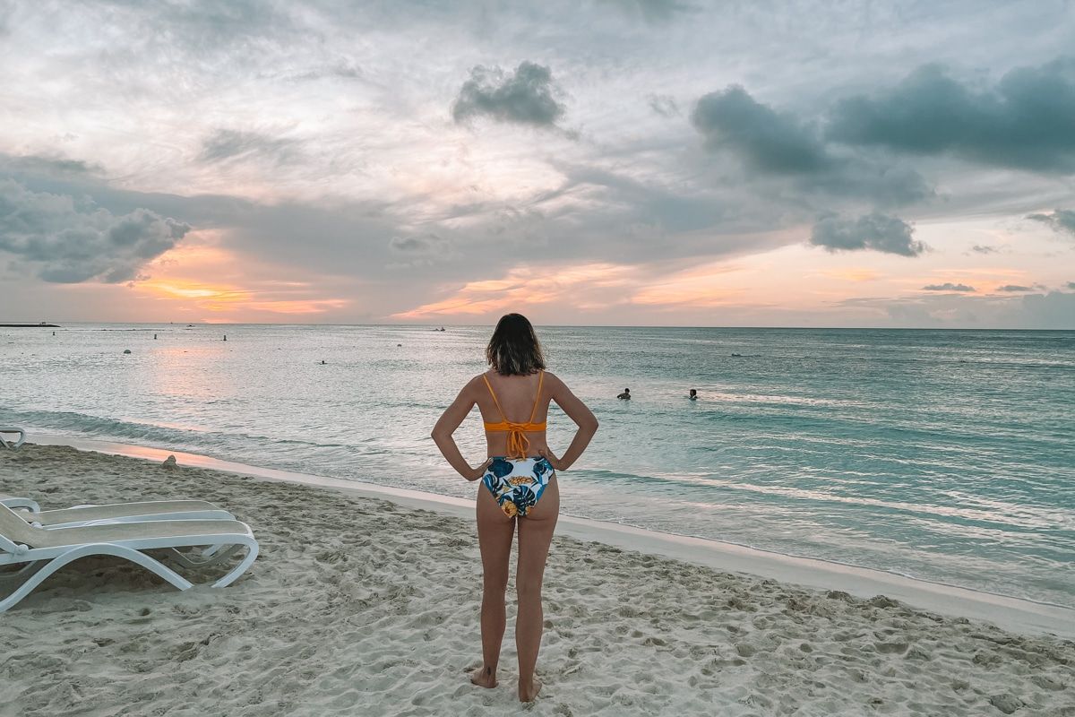 A woman in a yellow bikini top, and blue leaf-printed bottoms seen from behind walking on a tropical beach during a slightly overcast sunset.