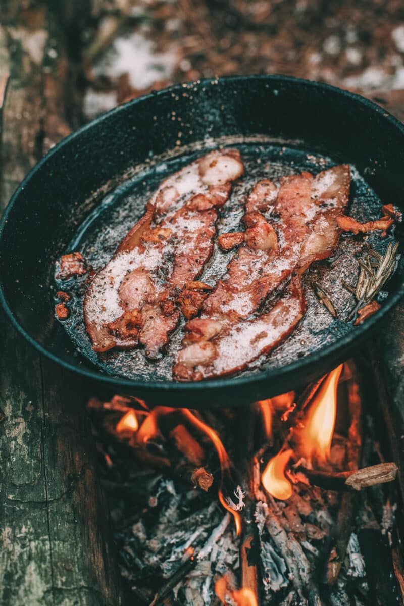 Cast iron skillets are some of the best campfire cooking kits, shown here over an open fire with two strips of bacon and a spring of rosemary sizzling inside the skillet.
