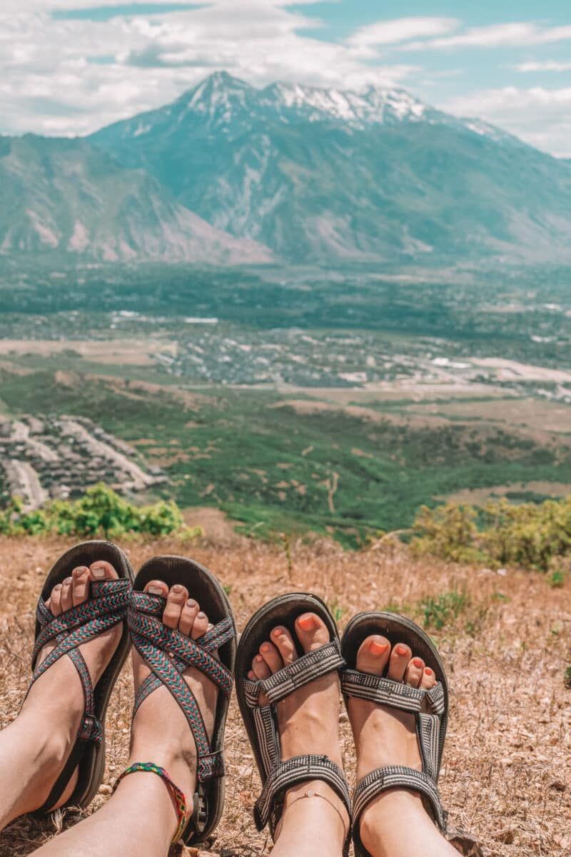 Some examples of the best minimalist hiking sandals on two pairs of women's feet at the top of a dry, grassy hill overlooking a valley and a mountain in the distance.