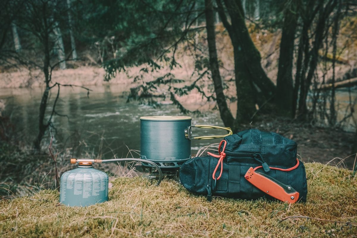 A camping cooking kit sitting on a mossy patch next to a black backpack with trees and a calm river in the background.