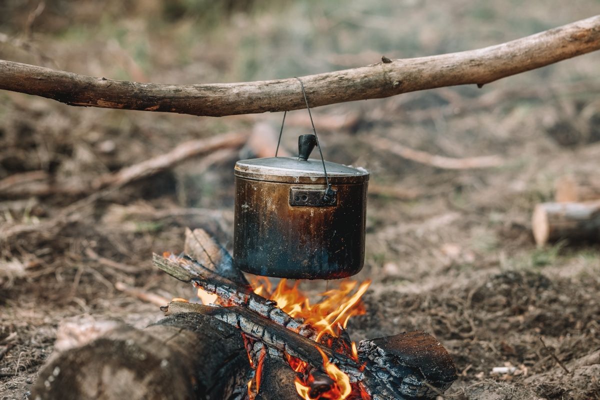 A blackened metal pot hanging from a branch over a campfire.