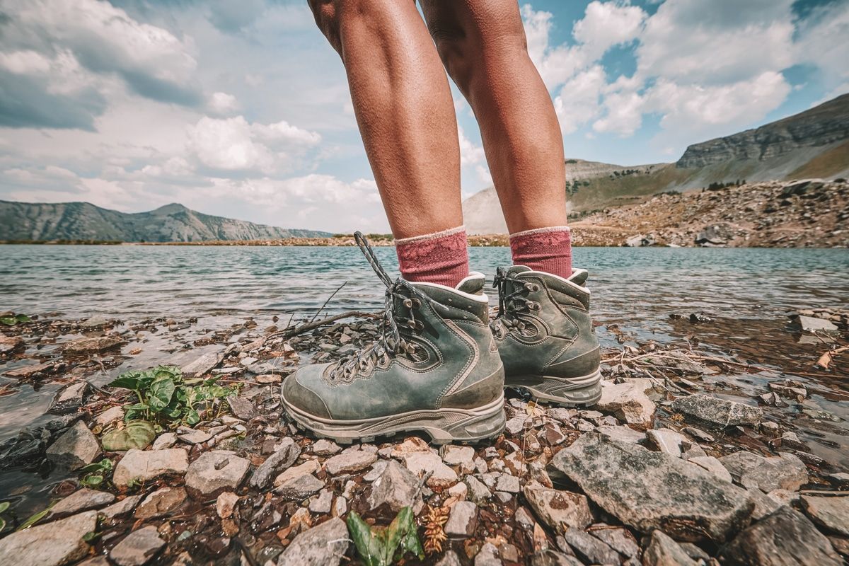 A pair of feet and muscular legs wearing blue hiking boots and maroon socks standing on a rocky lakeshore with mountains and partly-cloudy sky in the background.