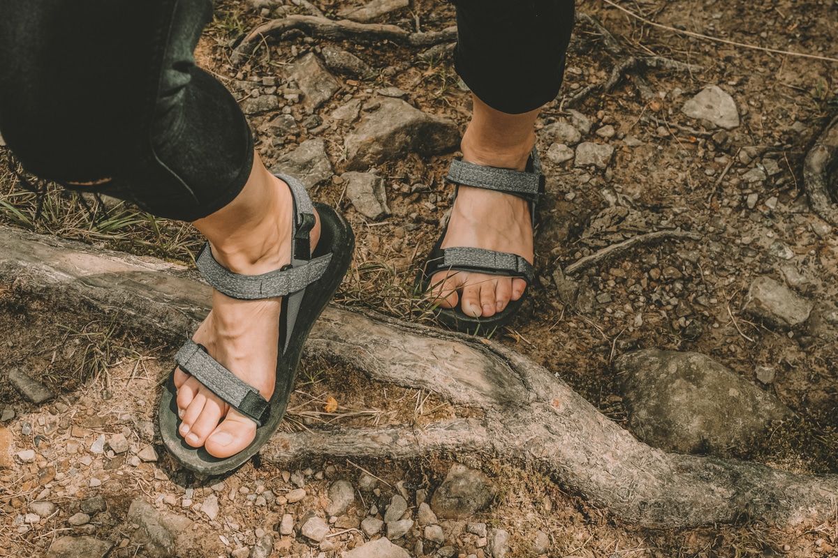 looking down at a pair of feet wearing grey hiking sandals while walking on a rocky dirt trail.