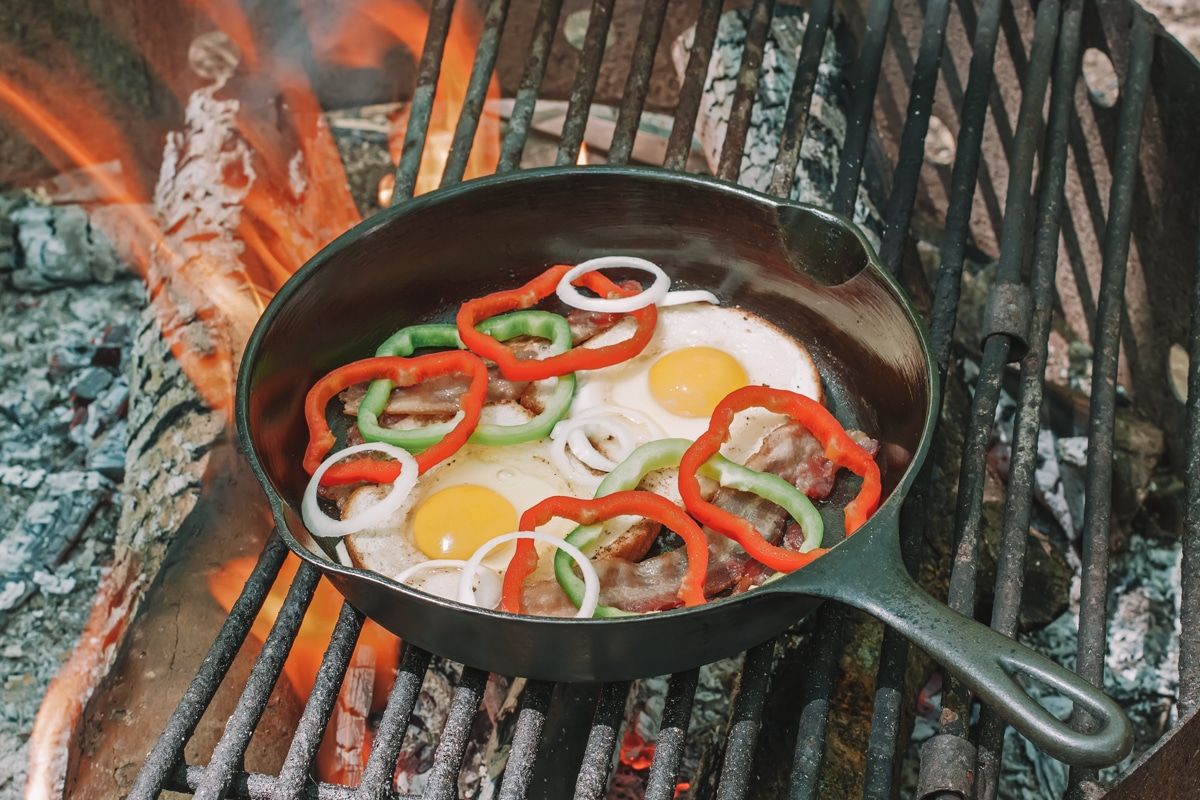 Two sunny-side-up eggs in a cast iron skillet with bacon and sliced bell peppers cooking on a grate over a campfire.