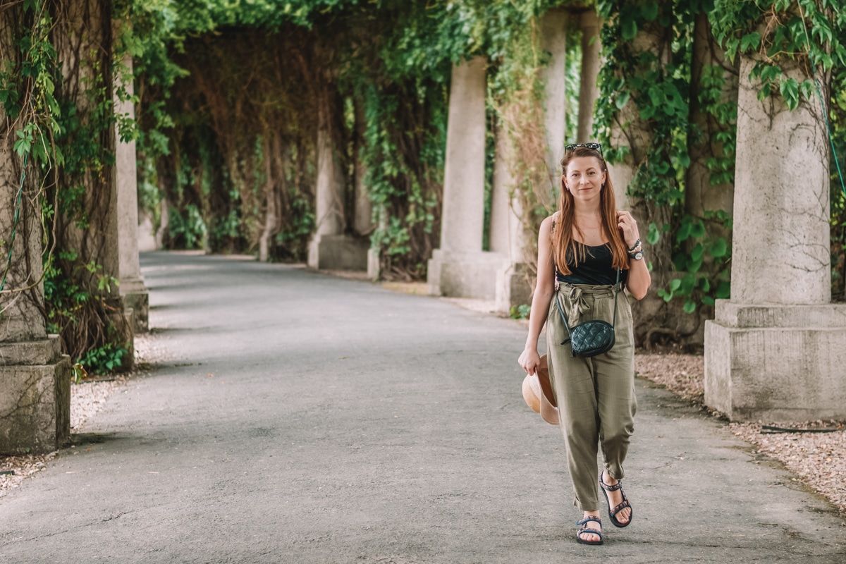 A young woman with long brown hair wearing a black top, olive green pants, and hiking sandals walks toward the camera on a paved path lined by ivy-covered stone columns.